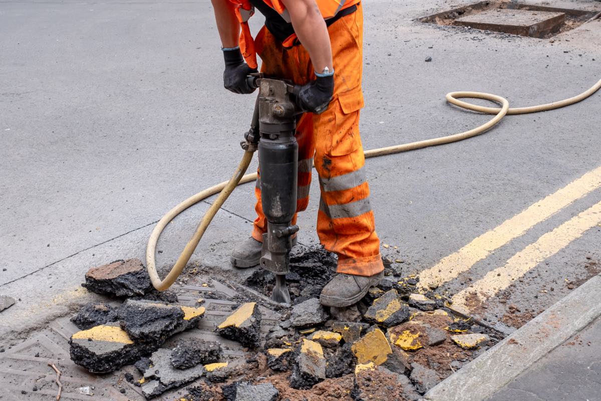 A road worker in hi-vis and gloves operating a pneumatic breaker on tarmac, the hand-arm vibration exposure that triggers Health Surveillance