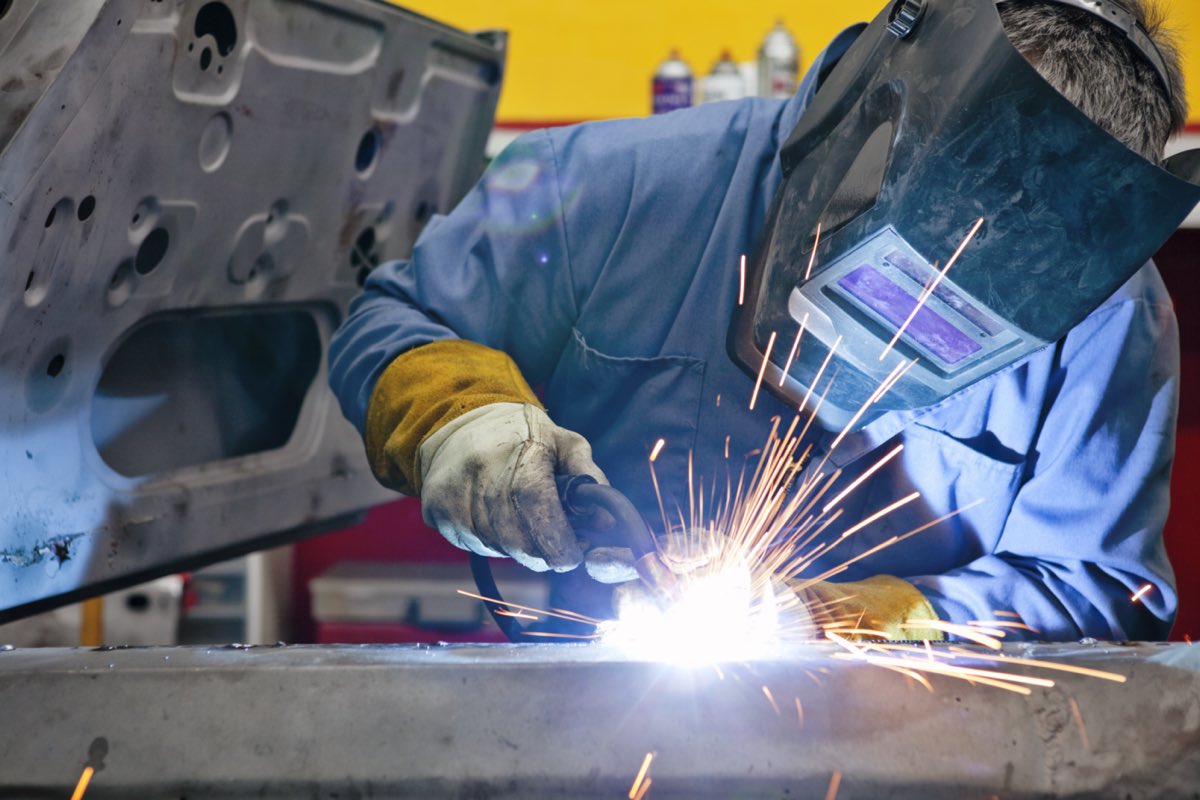 A welder in a flip-front welding helmet and leather gauntlets, MIG welding a steel panel, the fume and arc exposure a Health Surveillance programme covers